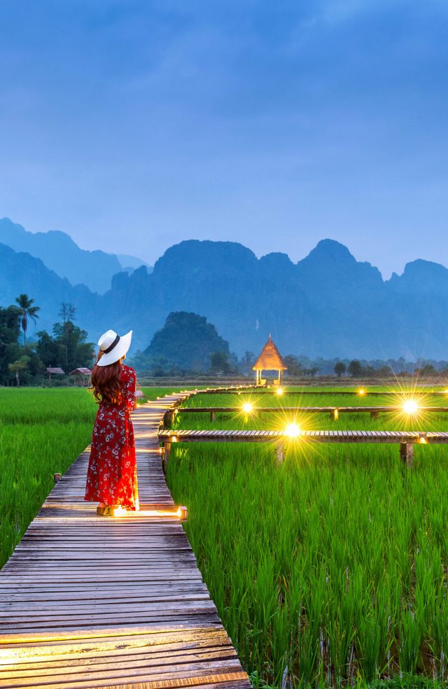 Young woman walking on wooden path with green rice field in Vang Vieng, Laos.
